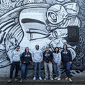AMCS former and current team wearing 'Marine' hoodies and other shop apparel, standing in front of street mural art of a face. 