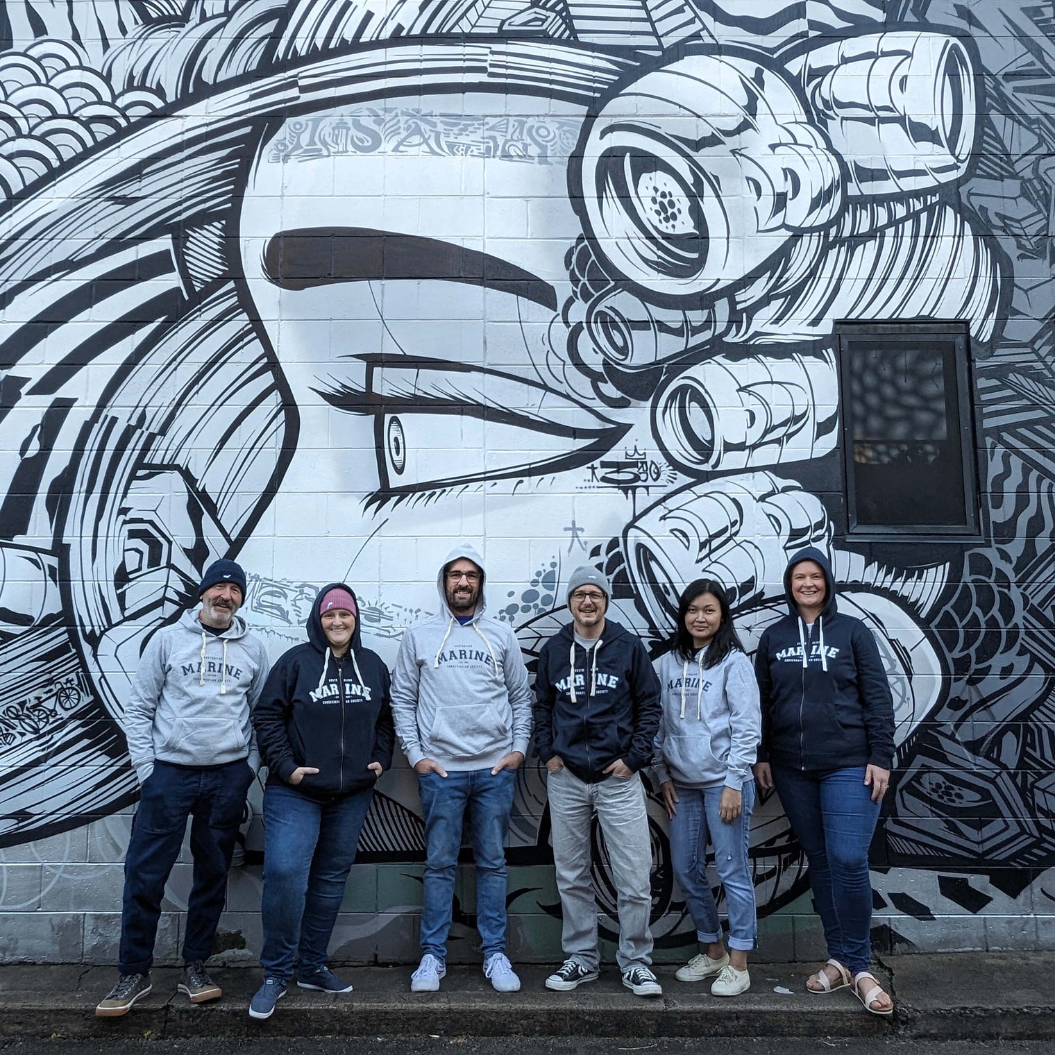 AMCS former and current team wearing 'Marine' hoodies and other shop apparel, standing in front of street mural art of a face. 