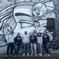 AMCS former and current team wearing 'Marine' hoodies and other shop apparel, standing in front of street mural art of a face. 